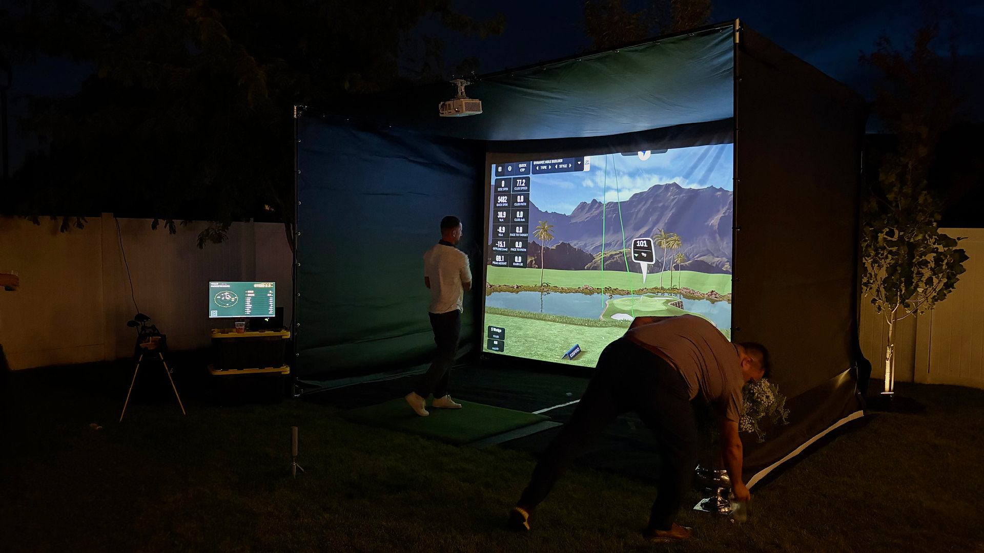 The TILT golf simulator set up in a backyard at night, with guests playing and the live standings on a monitor beside it